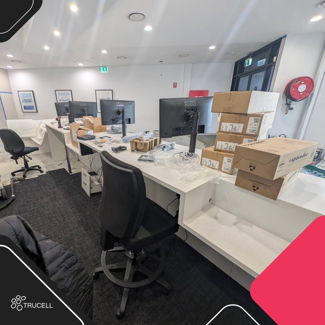 Estaciones de trabajo de recepción parcialmente instaladas en Medical Imaging Erina con dos monitores LG sobre brazos ajustables, cajas de hardware de HP y Spacedec preparadas para despliegue, pedestales con cajones en posición y soportes de CPU cableados bajo el mesón